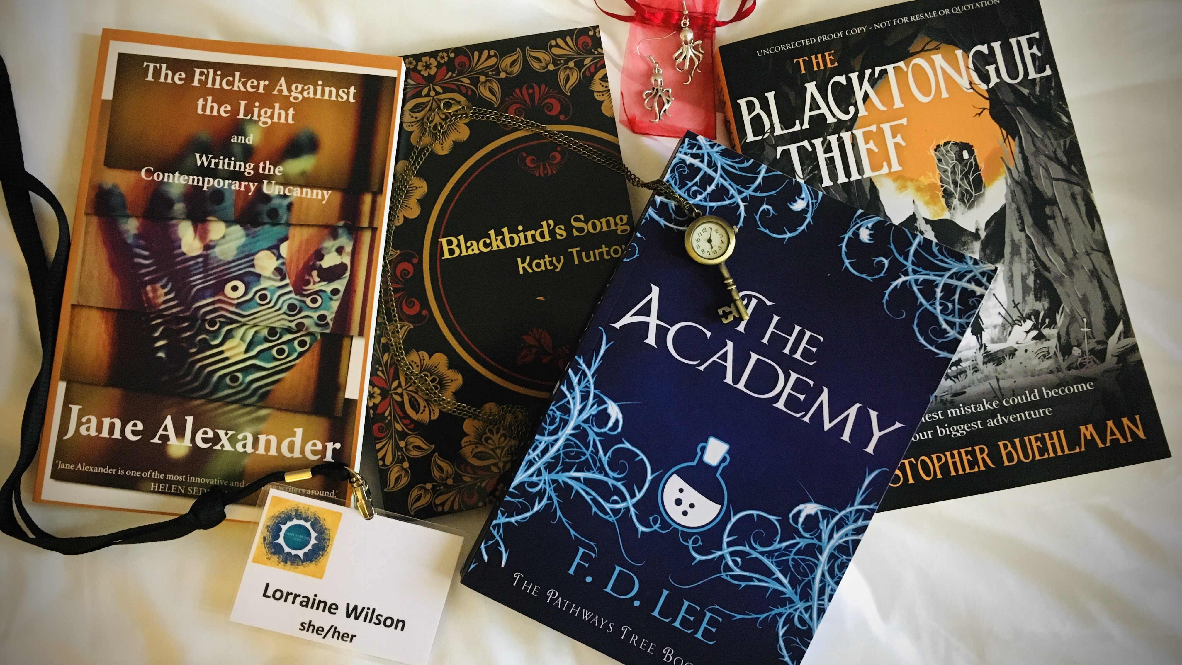 A photo showing four books, a necklace with clock pendant and a pair of octopus earrings. Books are The Blacktoungue Thief, Christopher Buehlman; The Academy, FD Lee; Blackbird's Song, Katy Turton, & The Flicker Against The Light, Jane Alexander.