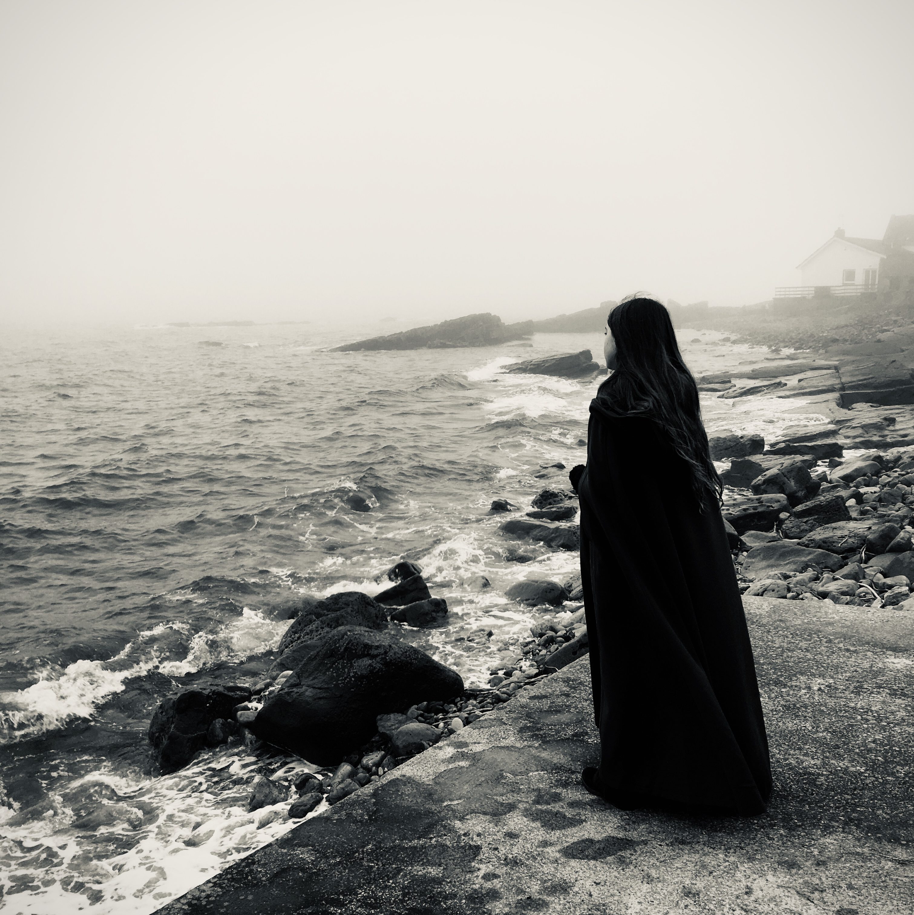 Black and white photo of a figure in a black cloak looking out to sea. 