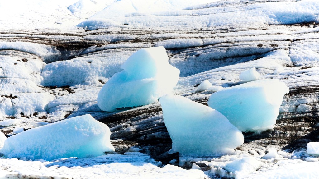 Close up photo of chunks of blue glacier ice resting on the glacier mass, which is white streaked with black lines. 