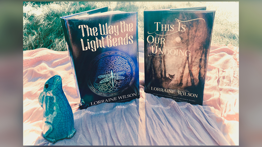 Photo of the hardbacks of both my books on a scarf on the lawn, backlit by sunlight & with a wee blue ceramic hare alongside. 