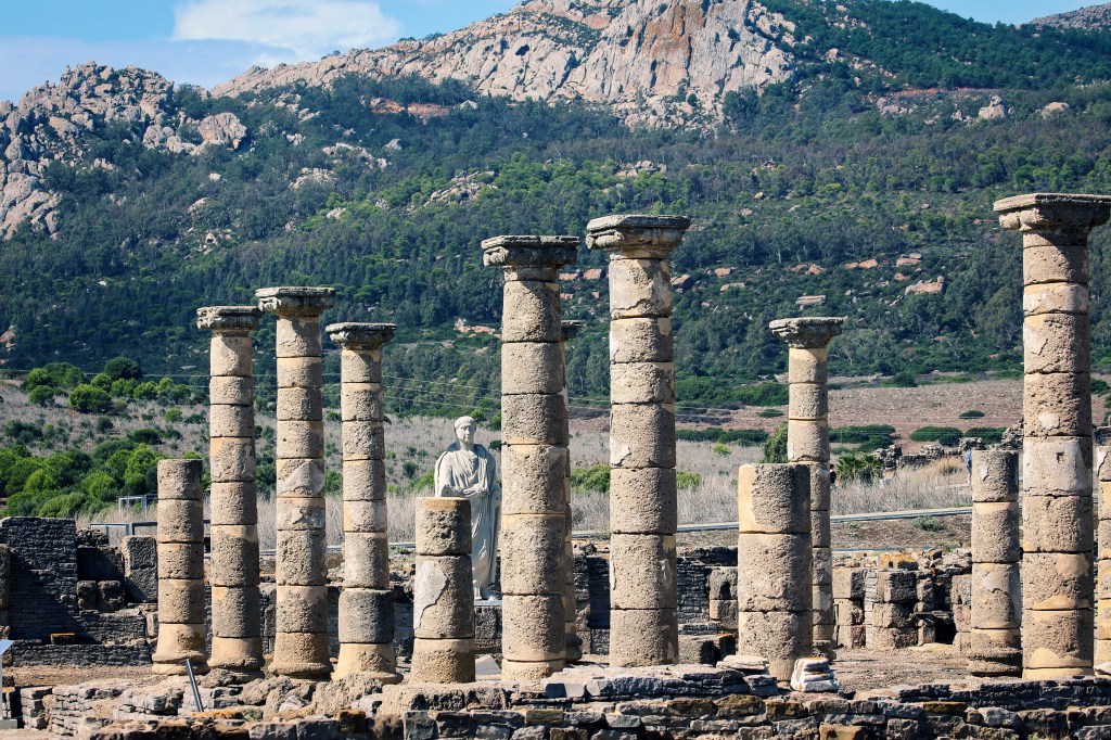 photo of standing columns of a grecian ruin on Cyprus, mountains in the background, the statue of some dude looking resigned and weary. 